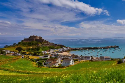 Blick über eine grüne Küstenlandschaft mit einer historischen Festung auf einem Hügel, einem kleinen Hafen mit zahlreichen Booten und einem Ort am Meer. Im Vordergrund liegen blühende Wiesen, darüber ein weiter blauer Himmel mit weißen Wolken.